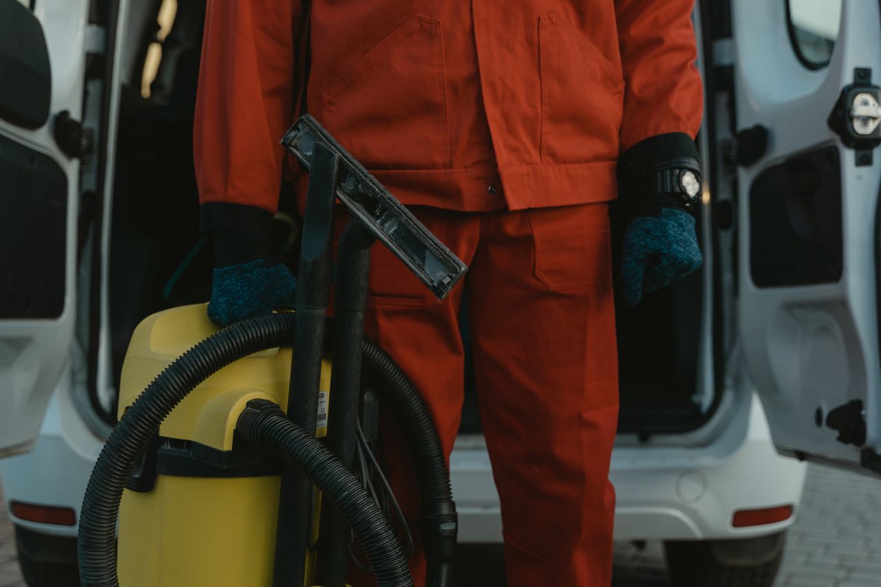 services-01 Une professionnelle du nettoyage en uniforme rouge tient un aspirateur près d’un van, prête à intervenir.